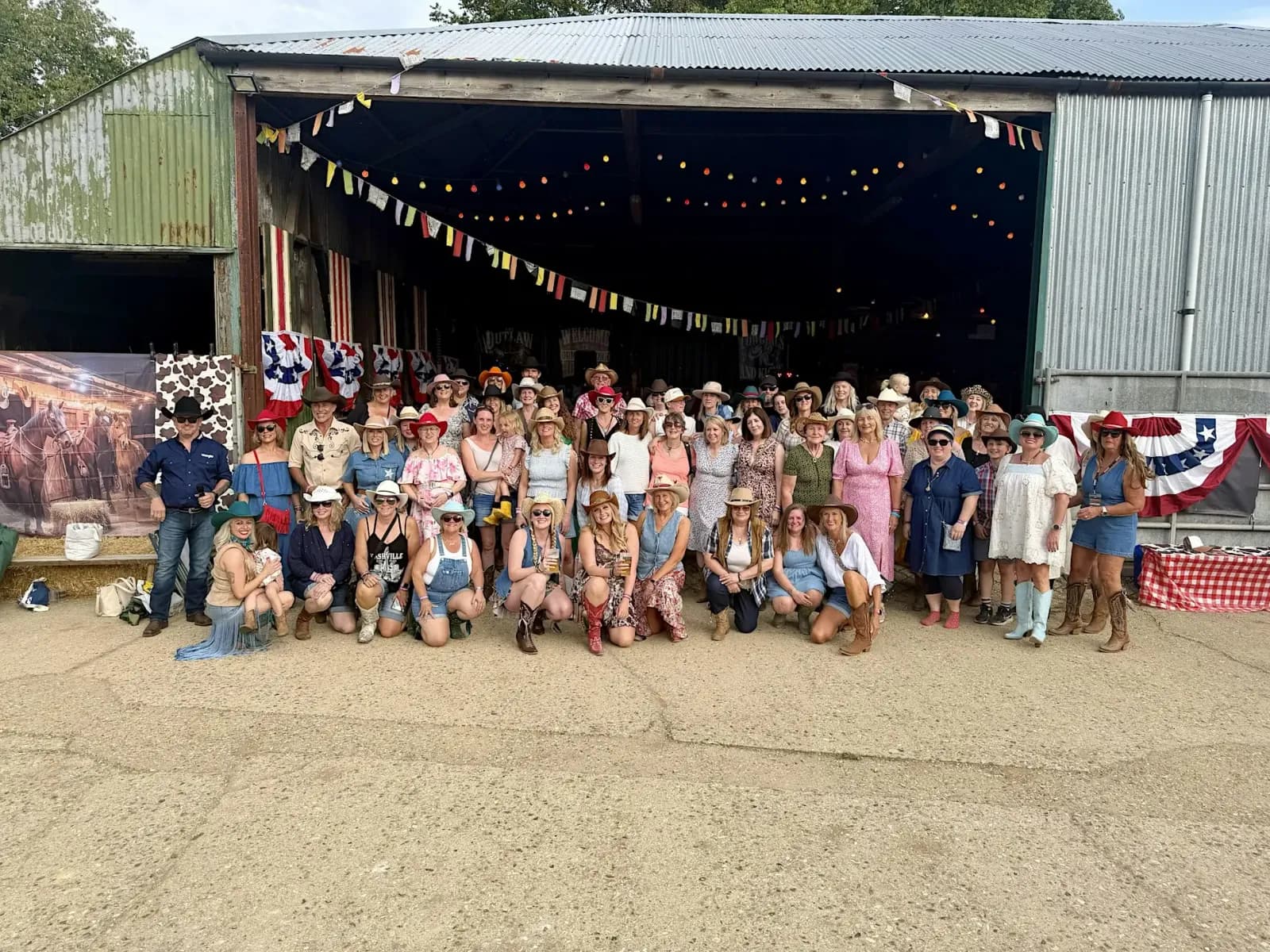 A large group of people, mostly women, dressed in cowboy-themed attire, pose together in front of a barn decorated with string lights and festive banners.