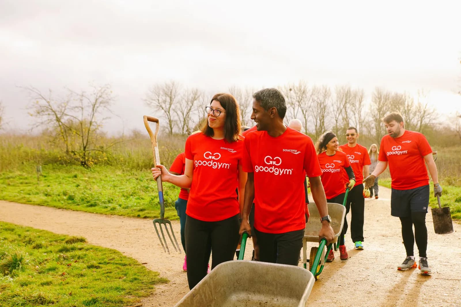 A group of people wearing red "Go Goodgym" shirts are walking on a dirt path, carrying gardening tools and wheelbarrows.