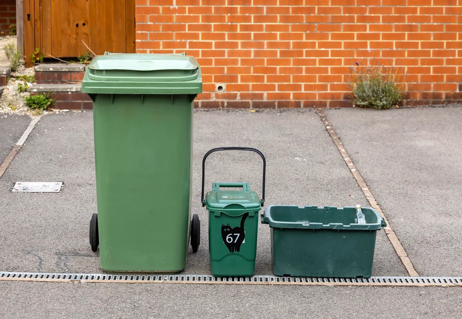 Three green recycling bins of different sizes are arranged on an asphalt driveway next to a red brick wall.