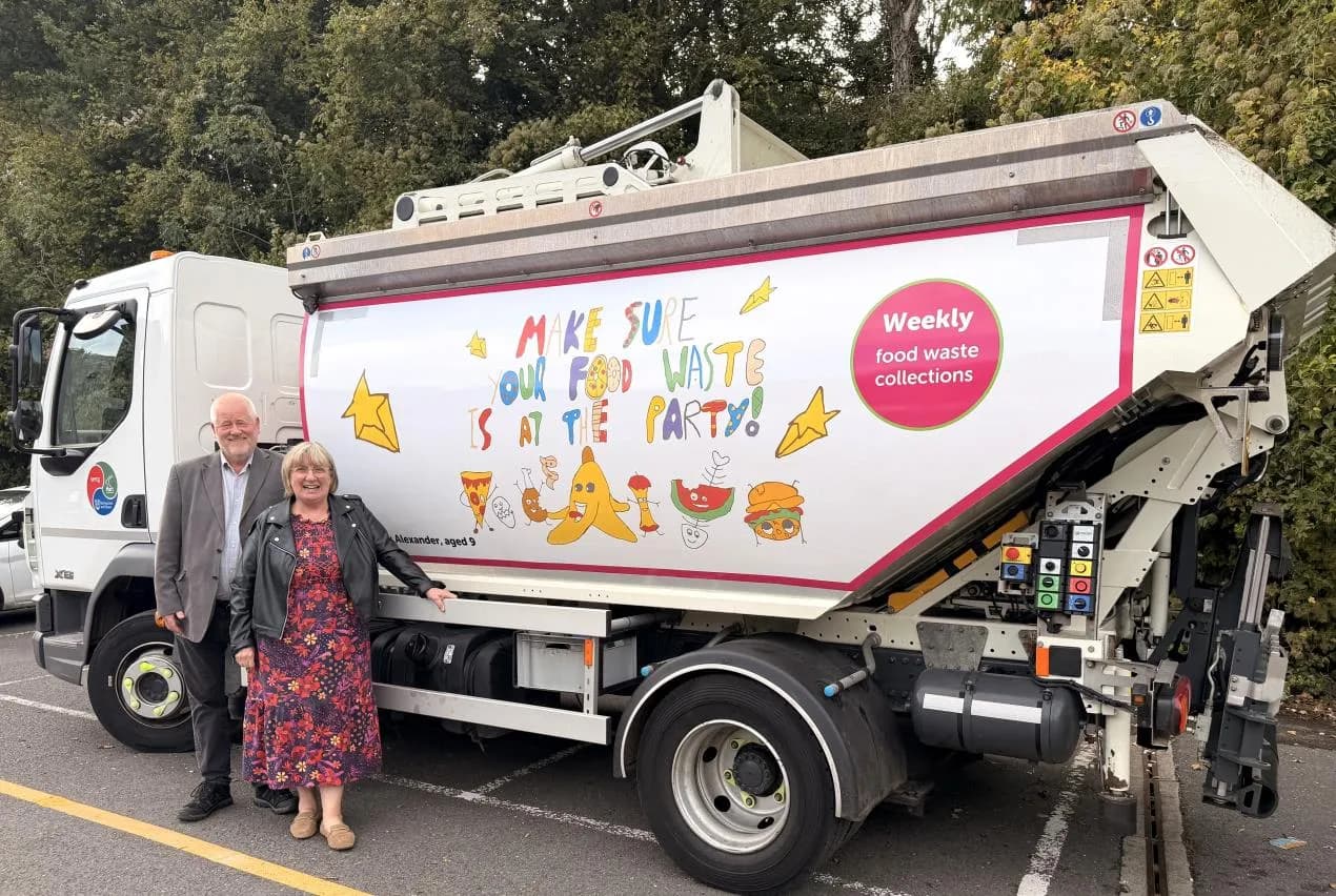 A man and a woman stand next to a white food waste collection truck decorated with cartoon food items and the text "Make sure your food waste is at the party!".